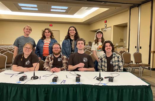 4 members of the quiz bowl team sit at a table ready to compete, with 4 supporting members standing directly behind them. 