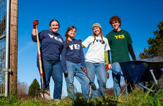 four students stand side-by-side smiling and holding shovels, wearing clothing with the Y.E.S. program logo on it.