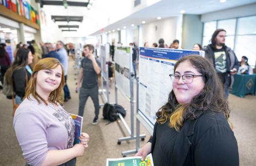 two students smiling as they stand in front of a poster presentation 