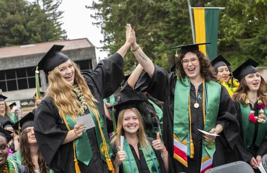 A photo of three graduates at commencement with two of them high fiving each other above the third graduate sitting down. . 