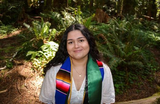 A photo of Carmen Benavides-Garb wearing graduation stoles with the forest in the background. 