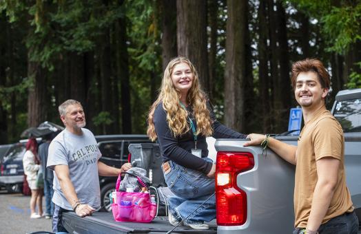 A student sits in the back of a flatbed truck full of her belongings. Two others are with the student - one is wearing a t-shirt that says "Cal Poly Humboldt Dad."