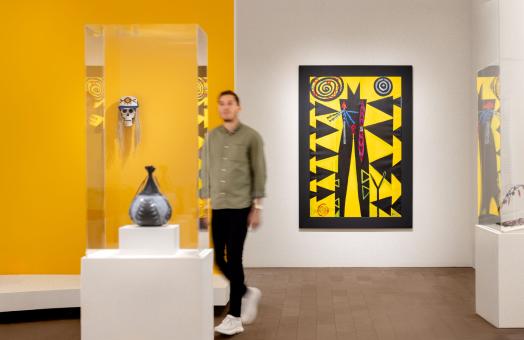 A photo of a blurred person walking through the de Young Museum. The photo features Indigenous artist Brian Tripp's "Someday You Might Have to Fight for What You Believe In" painting in the background. 