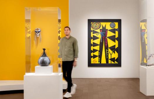 A photo of a blurred person walking through the de Young Museum. The photo features Indigenous artist Brian Tripp's "Someday You Might Have to Fight for What You Believe In" painting in the background. 