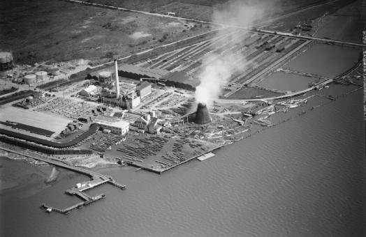 black and white aerial shot of an energy plant along the water, smoke bellowing out from a tower.