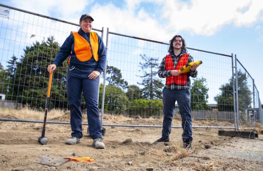two students standing conducting researching, one holding a sledgehammer and the other, a clipboard.