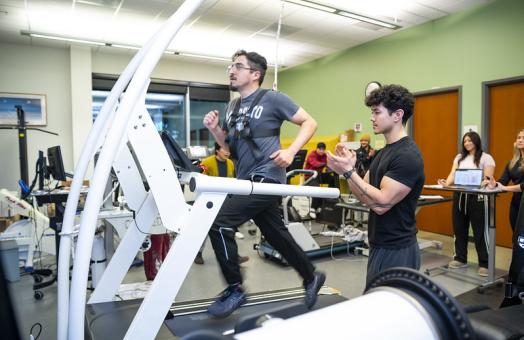 A photo of a student in the Kinesiology Human Performance Lab running on a treadmill wearing a vest that tracks their movement while another student cheers them on. 