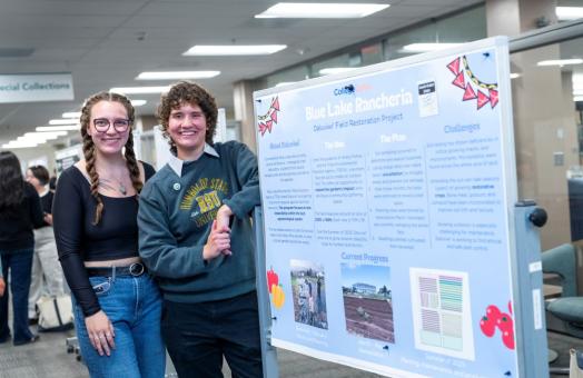 A photo of two students standing in front of their poster presentation in the Library during ideaFest in 2025. 