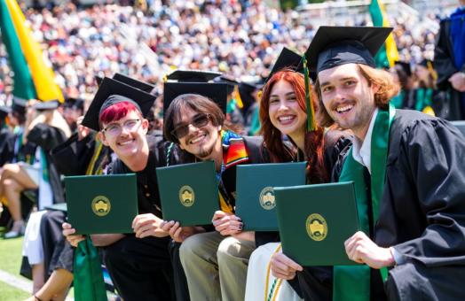 A photo of four graduates sitting down holding up their diploma covers on Commencement.