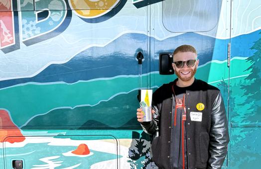 A photo of Carlo Solis standing in front of the mobile alumni center holding the Cal Poly Humboldt koozie during the Forever Humboldt at the San Francisco Giants game. 