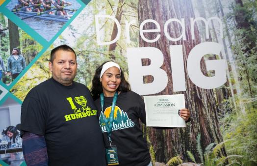 Parent and daughter standing in front of sign reading "Dream Big" holding an admittance letter. 
